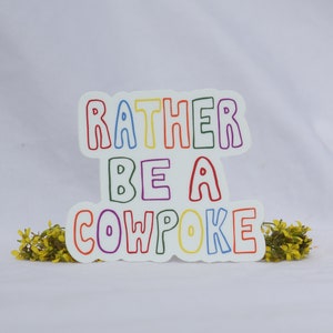 May include: A white sticker with the text "RATHER BE A COWPOKE" in colorful outlined letters. The sticker is decorated with yellow flowers on a white background.