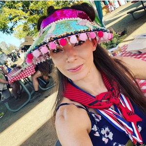May include: A woman wearing a colorful, sequined visor with pink pom-poms. The visor has a pink, blue, and green design. She is wearing a red and black striped scarf and a blue and white floral dress.