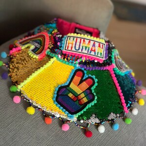May include: A colorful sequined collar with rainbow peace sign and "HUMAN" patch. The collar is decorated with pom-poms and beads.