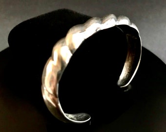 May include: A silver bracelet with a textured, wavy design. The bracelet is open and has a smooth, polished finish.