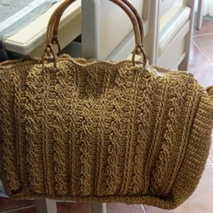 May include: A large, tan crocheted tote bag with a textured cable knit design. The bag features bamboo handles and a wide, open top. The bag is sitting on a light-colored chair.