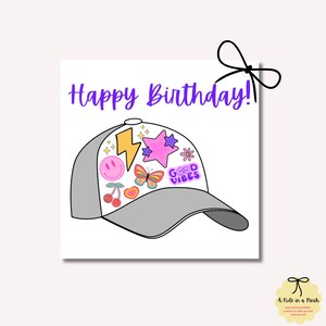 Könnte beinhalten: Eine weiße Karte mit dem Text "Happy Birthday!" in lila Schrift. Auf der Karte befindet sich eine graue Baseballkappe mit bunten Patches und Aufklebern. Die Patches beinhalten einen Blitz, einen Stern, ein Smiley, einen Schmetterling, Kirschen, ein Herz und ein Patch mit der Aufschrift "Good Vibes".