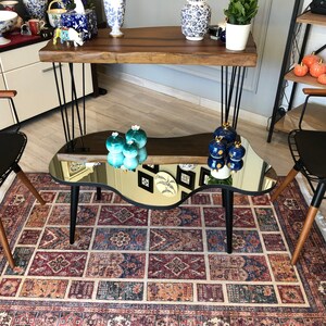 May include: A coffee table with a reflective, irregular-shaped mirror top and three black tapered legs. The table is positioned between two black wire-frame chairs with wooden armrests, all set upon a rectangular rug with a multi-colored geometric pattern.