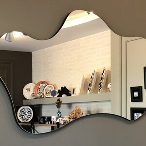 May include: A wall-mounted mirror with an irregular, organic, cloud-like shape and a thin black frame. The reflective surface shows a room interior with decorative plates, framed wall art, and various small objects displayed on a shelf.