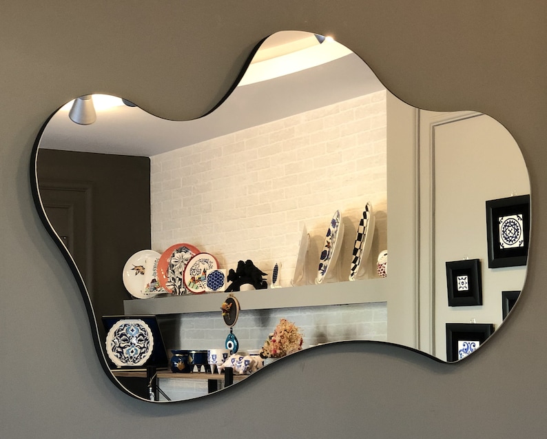 May include: A wall-mounted mirror with an irregular, organic, cloud-like shape and a thin black frame. The reflective surface shows a room interior with decorative plates, framed wall art, and various small objects displayed on a shelf.