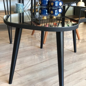 May include: A coffee table with a black frame and a mirrored top, featuring three tapered black legs. On the reflective surface sit three dark blue ceramic pomegranate-shaped ornaments with gold-colored crowns and gold-painted leaf details.