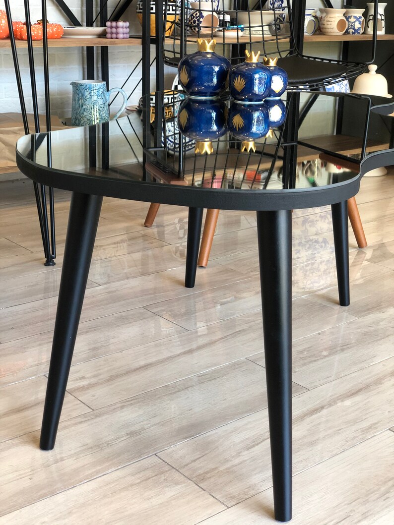 May include: A coffee table with a black frame and a mirrored top, featuring three tapered black legs. On the reflective surface sit three dark blue ceramic pomegranate-shaped ornaments with gold-colored crowns and gold-painted leaf details.