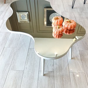 May include: An irregular, kidney-shaped coffee table with a reflective mirrored top surface and three white cylindrical legs. Two small, orange, white-spotted decorative pumpkins with gold stems are placed on the mirrored surface.