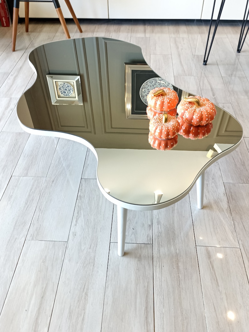 May include: An irregular, kidney-shaped coffee table with a reflective mirrored top surface and three white cylindrical legs. Two small, orange, white-spotted decorative pumpkins with gold stems are placed on the mirrored surface.