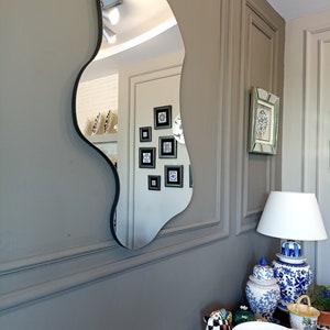 May include: A wall-mounted mirror with an irregular, organic, wavy shape and a thin black frame. The reflective surface shows a room interior with framed wall art, a lamp, and various decorative ceramic objects on a surface below.