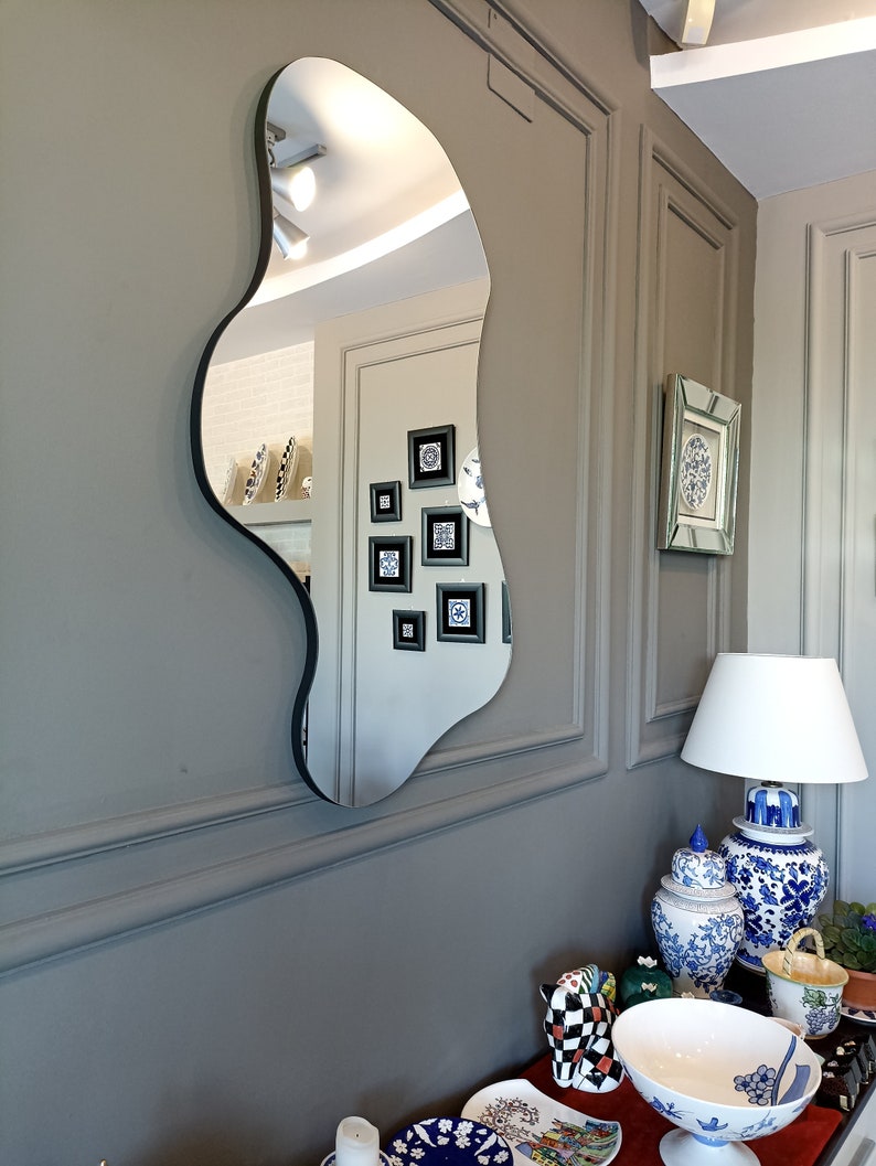 May include: A wall-mounted mirror with an irregular, organic, wavy shape and a thin black frame. The reflective surface shows a room interior with framed wall art, a lamp, and various decorative ceramic objects on a surface below.