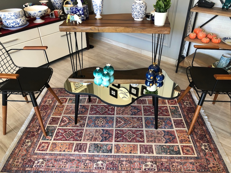 May include: A coffee table with a reflective, irregular-shaped mirror top and three black tapered legs. The table is positioned between two black wire-frame chairs with wooden armrests, all set upon a rectangular rug with a multi-colored geometric pattern.