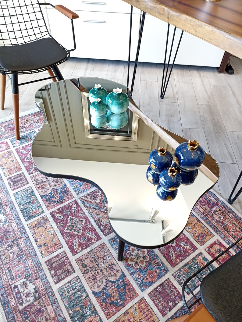 May include: An irregular, kidney-shaped coffee table with a reflective mirrored surface and a black edge. The table features dark wooden legs and displays several decorative ceramic pomegranate-shaped ornaments in shades of blue and teal.