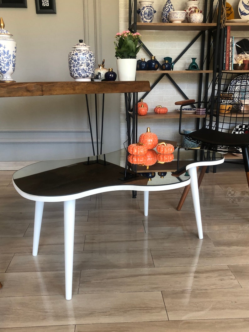 May include: A kidney-shaped coffee table with a reflective mirrored top and three tapered white legs. Three small orange decorative pumpkins with gold stems are placed on the surface, reflecting in the mirror finish of the table top.