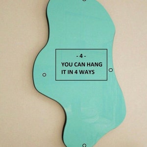May include: An irregular, organic-shaped light teal mirror featuring four small circular mounting holes positioned along its perimeter. A rectangular black border in the center contains the text: - 4 - YOU CAN HANG IT IN 4 WAYS.