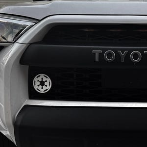 May include: A white Toyota SUV with a black grille and the word "TOYOTA" in silver lettering. The grille has a black and white Star Wars Imperial symbol emblem.
