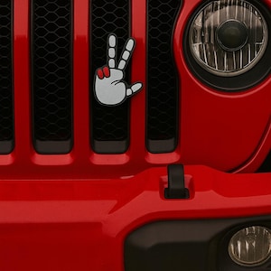 Peut inclure: Gros plan sur une calandre de Jeep rouge avec un emblème de signe de paix. L'emblème est une main blanche avec des accents rouges. Le mot "Jeep" est visible en noir au-dessus de la calandre.