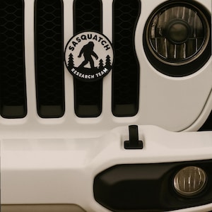 May include: Close-up of a white Jeep grill with black accents. A circular Sasquatch Research Team emblem is centered on the grill. The word "Jeep" is above the grill in black lettering. The vehicle's headlight and fog light are visible.