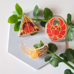 Può includere: Tre spille in resina con design a tema Harry Potter. La prima spilla è di forma triangolare con un design rosso e oro di una fenice. La seconda spilla è di forma rotonda con un design rosso e oro di una fenice con le ali spiegate. La terza spilla è un design dorato di un boccino d'oro con le ali spiegate.