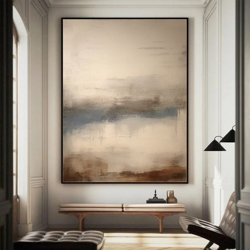 May include: Abstract painting with a beige background and brown, blue, and gray brushstrokes. The painting is framed in a black frame and is hanging on a white wall.