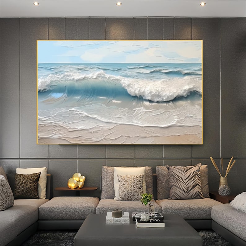 May include: A framed ocean wave painting. The artwork depicts a breaking wave in shades of blue and white, with a sandy beach in the foreground. The painting is in a gold frame and hangs above a gray sectional sofa.