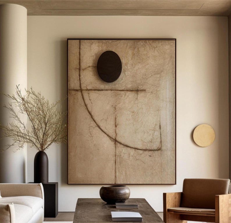 Puede incluir: Una gran pintura abstracta con un fondo marr&oacute;n y un c&iacute;rculo negro en el centro. La pintura est&aacute; enmarcada en negro y cuelga de una pared beige. Hay una peque&ntilde;a decoraci&oacute;n de pared redonda de color dorado a la derecha de la pintura.