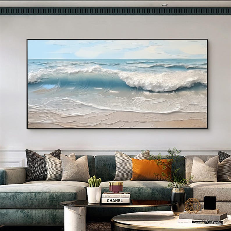 May include: A framed ocean wave painting. The artwork depicts a breaking wave in shades of blue and white, with a sandy beach in the foreground. The painting is displayed above a sofa with decorative pillows.