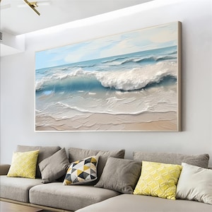 May include: A large framed painting depicts a textured ocean scene. The artwork features a light blue sky, turquoise water, and white foamy waves crashing onto a sandy beach. The painting is displayed above a gray sofa with decorative pillows.