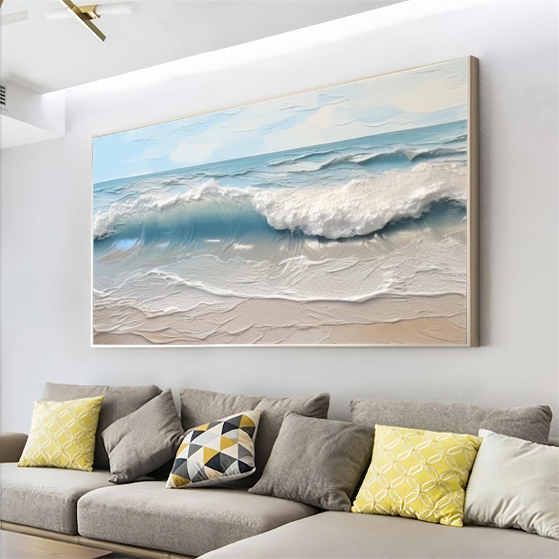 May include: A large framed painting depicts a textured ocean scene. The artwork features a light blue sky, turquoise water, and white foamy waves crashing onto a sandy beach. The painting is displayed above a gray sofa with decorative pillows.