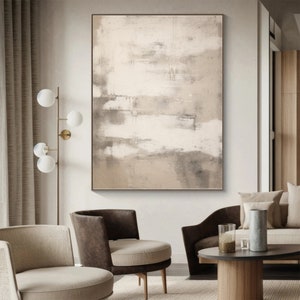 May include: Abstract painting with shades of beige, brown, and white. The painting is framed and hanging on a white wall in a living room setting.