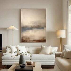 May include: Abstract painting with brown, beige, and blue hues. The painting depicts a landscape with a horizon line and a reflection of the sky in water. The painting is framed and hanging on a wall in a living room.