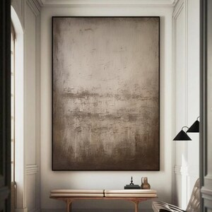 May include: A large abstract painting in shades of brown and beige hangs on a white wall in a modern interior. The painting has a textured surface and a subtle gradient from light to dark.
