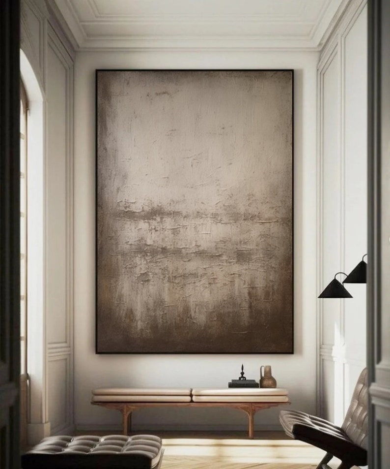 May include: A large abstract painting in shades of brown and beige hangs on a white wall in a modern interior. The painting has a textured surface and a subtle gradient from light to dark.