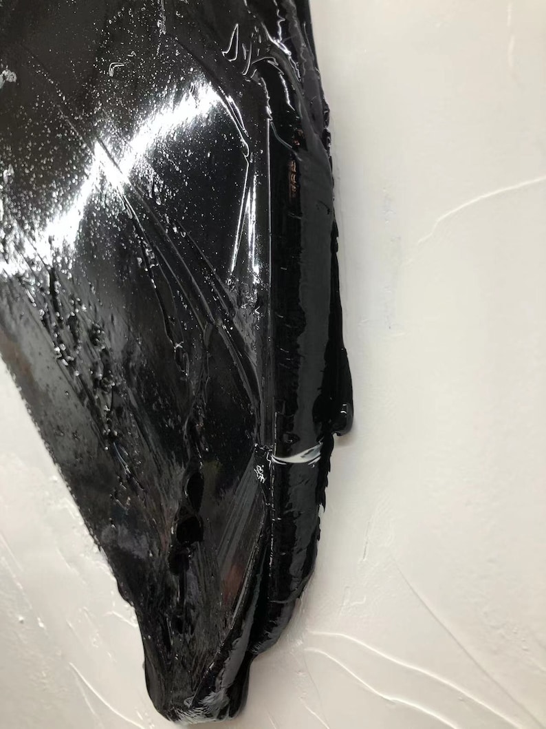 May include: A close-up of a black, abstract, textured, three-dimensional art piece. The piece is made of a thick, glossy material and has a smooth, flowing surface. The piece is mounted on a white wall.