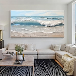 May include: Large rectangular canvas art depicting an ocean scene. The artwork features a light blue sky, waves in shades of blue and white, and a sandy beach. The painting is displayed above a modern living room sofa.