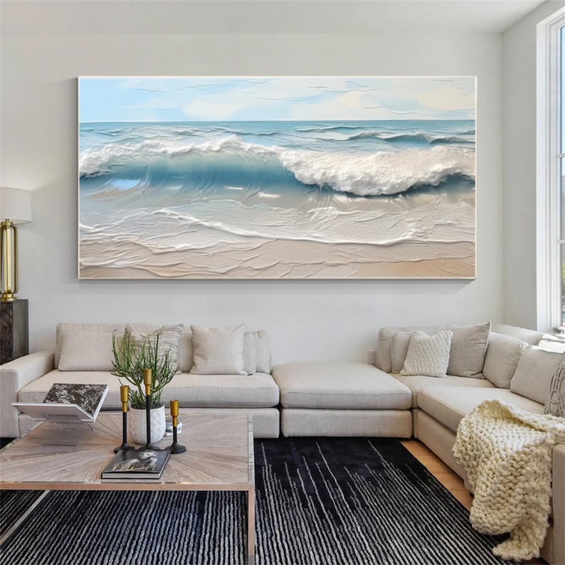 May include: Large rectangular canvas art depicting an ocean scene. The artwork features a light blue sky, waves in shades of blue and white, and a sandy beach. The painting is displayed above a modern living room sofa.