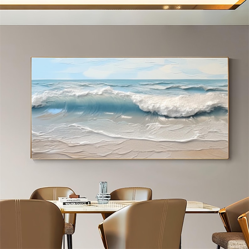 May include: A framed painting depicts a seascape with a textured, three-dimensional effect. The artwork showcases a dynamic ocean scene with foamy white waves crashing onto a sandy beach under a light blue sky. The painting is encased in a gold-colored frame.