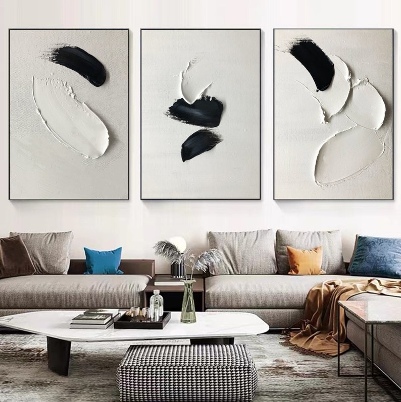 May include: Three abstract paintings in black and white with thick brushstrokes and textured paint. The paintings are hung on a white wall above a gray couch with pillows and a coffee table.