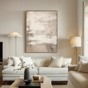 May include: A white couch with beige throw pillows sits in a living room with a large abstract painting in shades of brown and white hanging on the wall. Two lamps with white shades stand on either side of the couch.