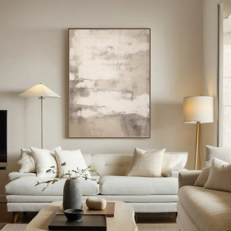 May include: A white couch with beige throw pillows sits in a living room with a large abstract painting in shades of brown and white hanging on the wall. Two lamps with white shades stand on either side of the couch.
