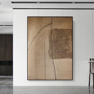 May include: Abstract art piece in a black frame. The artwork features a textured, neutral color palette with shades of beige and brown. Dark, linear elements create a contrast against the lighter background, adding depth and visual interest to the composition.