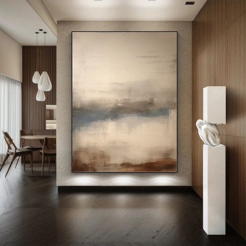 May include: Abstract painting in shades of brown, beige, and blue, hanging on a wall in a modern interior. The painting is framed in black and has a textured surface.