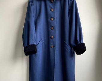 May include: A long, blue coat with a black velvet collar and cuffs. The coat has a button-up front and two pockets. The buttons are brown. The coat is hanging against a white wall.