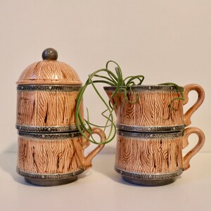May include: A set of four ceramic mugs with a wood grain design and metal accents. The mugs are stacked and have a lid. The mugs are brown and beige with a gray metal band around the top.