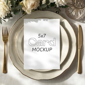 May include: A white 5x7 card mockup featuring the text 5x7 Card MOCKUP in black outline and solid lettering, centered on a white plate with a gold rim, accompanied by a gold fork and a gold knife, all arranged on a white tablecloth.