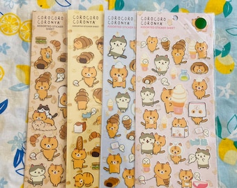 May include: Four sticker sheets with the text "COROCORO CORONYA ASSORTED STICKER SHEET". Each sheet features different illustrations of cartoon cats with food items like sandwiches, croissants, and ice cream. The background is a blue and white floral pattern.