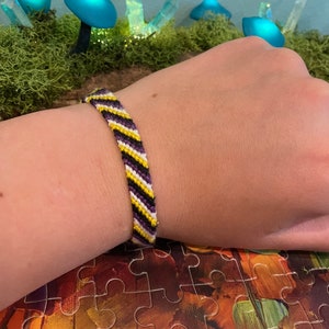 May include: A friendship bracelet with yellow, white, and purple stripes. The bracelet is worn on a person's wrist.