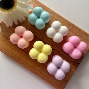 Puede incluir: Seis bombas de baño en forma de flor de color pastel sobre una tabla de cortar de madera. Las bombas de baño son en tonos de azul, blanco, rosa, amarillo y morado.