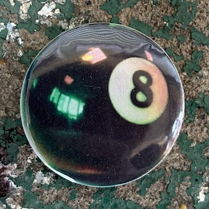 May include: A circular pin-back button featuring a black billiard ball design with a white circular inset containing the black number 8. The surface has a glossy finish with light reflections and a subtle iridescent sheen across the dark background.