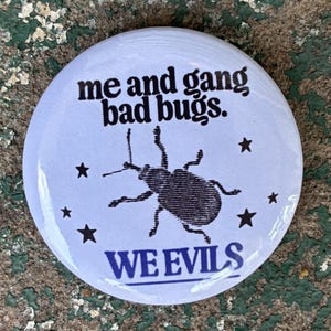May include: A circular pinback button with a light purple background. It features a black graphic of a beetle surrounded by small black stars. The text reads me and gang bad bugs. in black and WE EVILS in purple at the bottom with a line.
